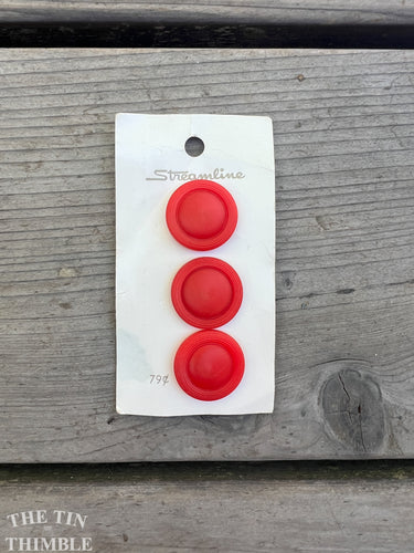 A set of three red plastic buttons on their original button card against a wood background.