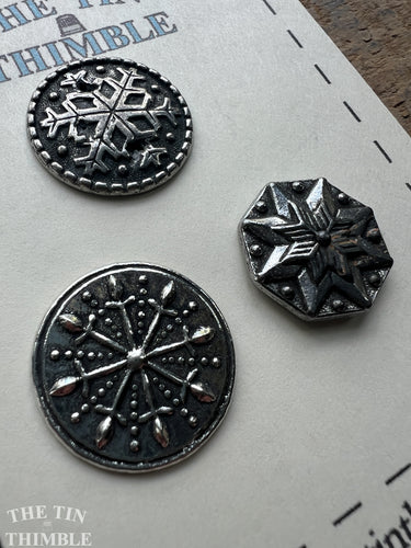 Three Scandinavian silver buttons with star and snowflake designs on a textured surface.